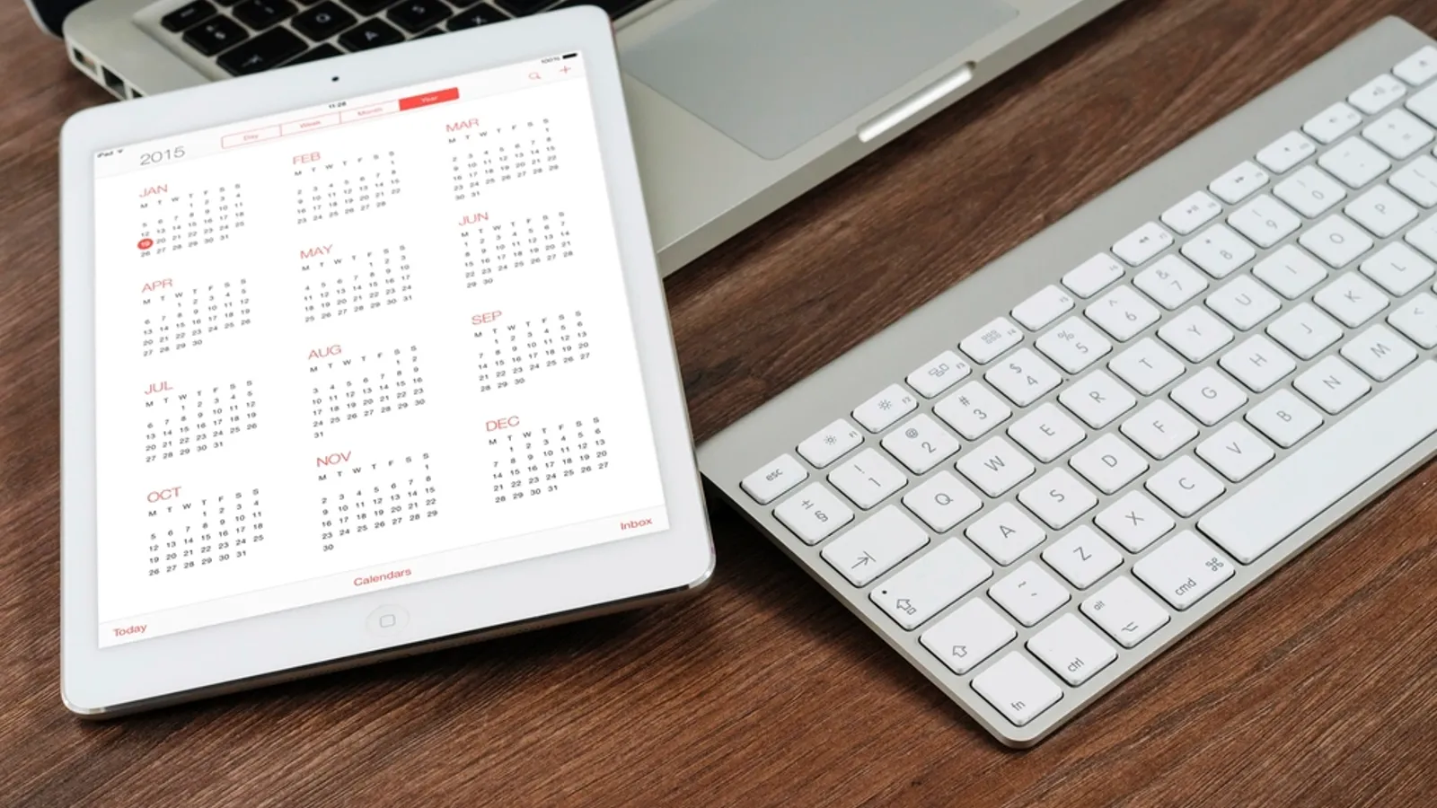 A tablet showing a monthly calendar sits beside a keyboard on a wooden desk, ready for booking meetings.