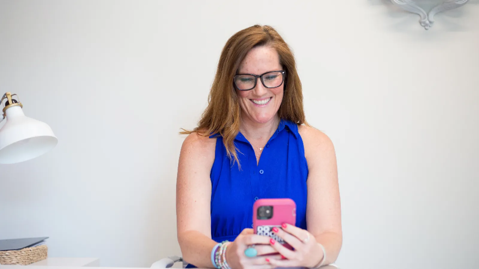 A smiling saleswoman in a blue top holds her phone at a bright desk.