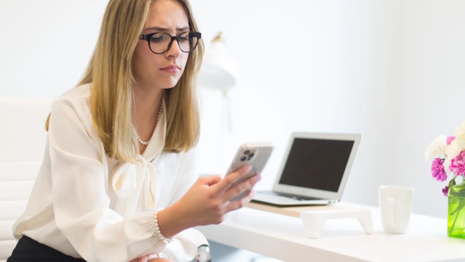 A woman at a desk looks down at her phone with a laptop open beside her, getting ready for an outbound sales call.